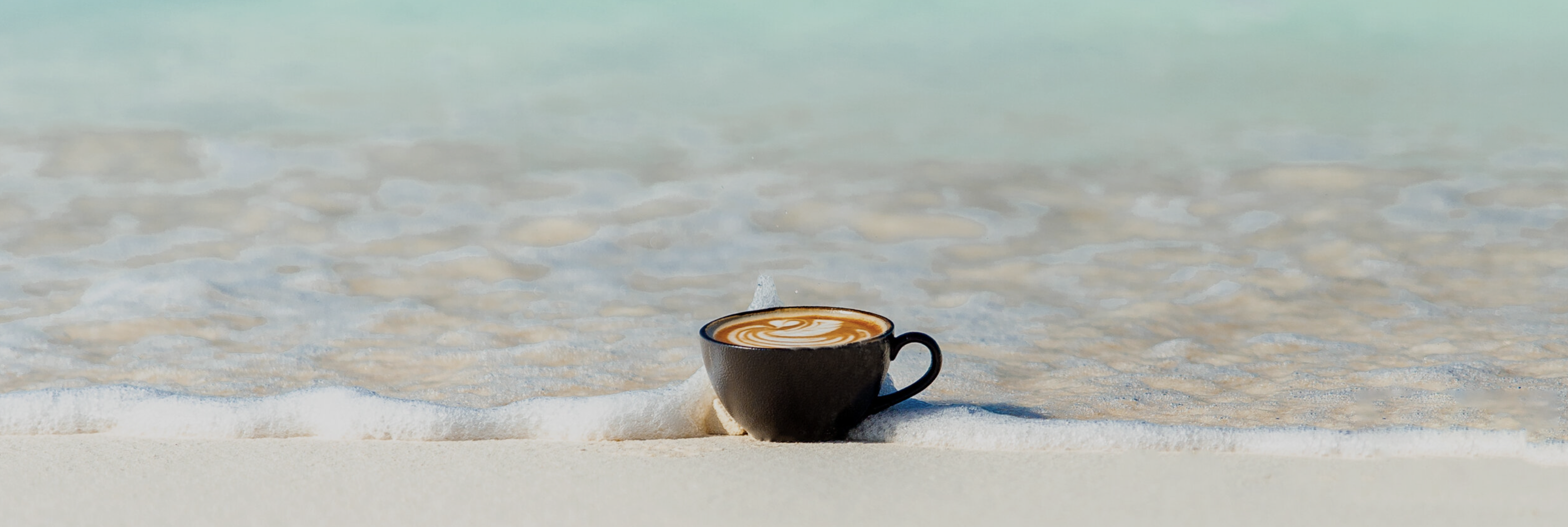 A cup of capuccino in the beach touching the water, proposing that Florida and fresh brewed coffee from roasted beans is a mperfect match!